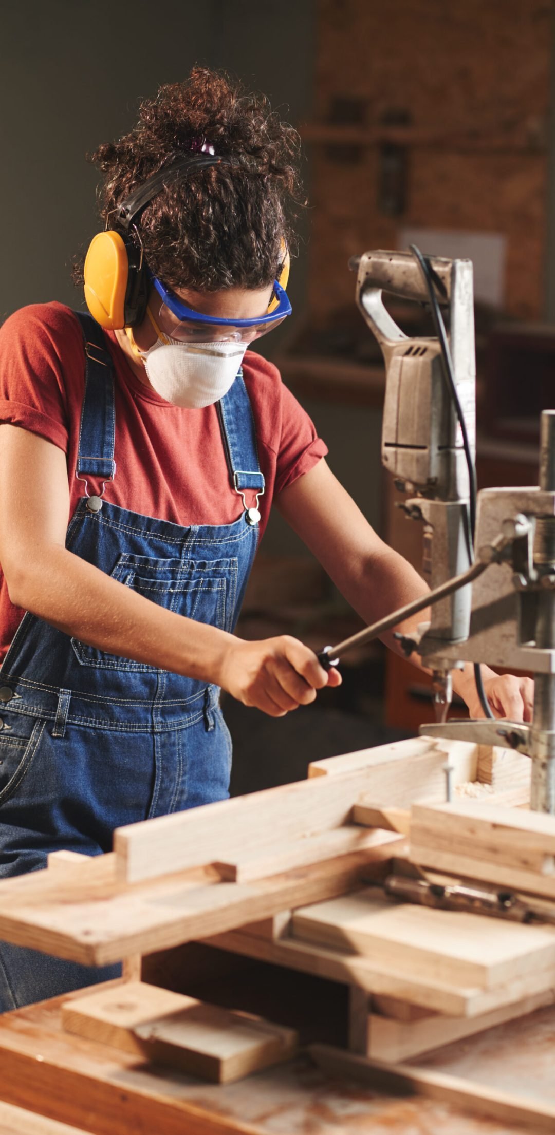 Young female carpenter in protective eyewear and ear defenders pressing lever on woodworking machine while cutting wooden planks